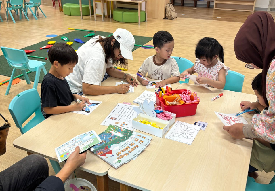 Adults and children sit at a classroom table coloring worksheets with markers, scissors, and supplies.